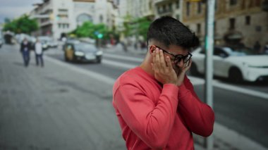Gözlüklü genç adam hareketli bir şehir arka planında duygularını ifade ediyor. Aydınlık bir günde taksiler ve yayalar geçiyor..