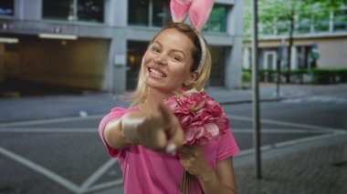 Pembe tişörtlü kadın tavşan kulağı takıyor, parmaklarını gösterip sokakta buket tutuyor..