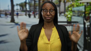 Kıvırcık saçlı, gözlüklü ve sarı tişörtlü bir kadın güneşli bir sahil gezisinde sürpriz yapıyor. Arka planda palmiye ağaçları, canlı ve canlı bir atmosfer var..