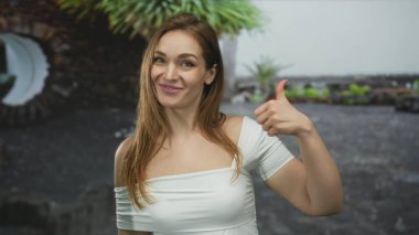 Beyaz omuzlu bir kadın açık hava binasının önünde baş parmak hareketi gösteriyor. Onaylama iyimserliği.