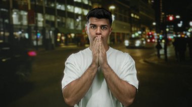 Genç bir adam gecenin bir yarısı şehir caddesinde parlak ışıklar ve bulanık şehir geçmişiyle çevrili endişeli bir ifadeyle beyaz bir gömlek giyerek düşünceye dalıyor..