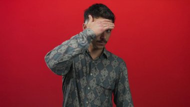 Stüdyodaki izole edilmiş kırmızı arka plan duvarına karşı elini yüzünü kapatan İspanyol genç adam..