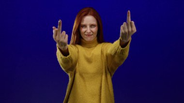 Sarı kazaklı bir kadın, mavi bir arka plana karşı saldırgan bir el hareketi sergiliyor. Küstah bir ifadeyle, kırmızı saçlar, canlı mavi duvara karşı duruyor. Asi havasını arttırıyor..