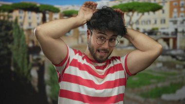 Kırmızı çizgili tişörtlü, sakallı genç İspanyol adam güneşli Roma harabelerinde kulak hareketi yaparak antik mimari ortamında neşeli ve güneşli bir anı yakalıyor..