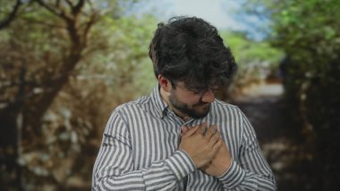 Sakallı genç bir adam parkta göğsünü sıkıyor, muhtemelen yeşil ağaçlarla çevrili kalp rahatsızlığı yaşıyor ve sıkıntılı bir gün ışığı görüyor..