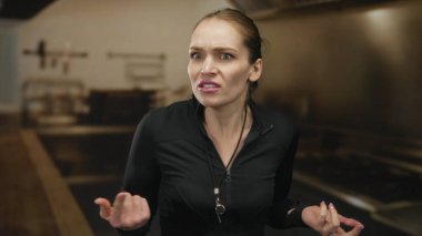 Arka planda bulanık ağırlıklarla stüdyoda siyah ceketli, ıslıkla parmaklarını göğsüne gösteren bir kadın antrenör..