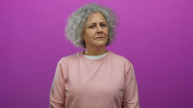 Kıvırcık gri saçlı, pembe bluzlu, hayat dolu pembe arka planda duran ve merak ve tefekküre dalmış bir kadın..