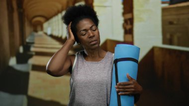 Antik üniversite koridorunda mavi yoga minderi tutan kadın gözleri kapalı, tarihi bir iç mekanda farkındalık ve sükuneti somutlaştırıyor..