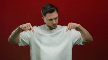 Sakallı, beyaz gömlekli genç adam canlı kırmızı arka plana karşı aşağı doğru işaret ediyor, izole stüdyo ortamında fikir ya da yön aktarıyor kendine güvenen bir tavırla..