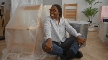 Yeni dairede gülümseyen Afrikalı bir kadın. Paketlenmemiş mobilya ve bitkilerle yerde oturuyor. Günlük giysiler giyiyor ve mutlu görünüyor..