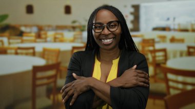 Kolları çapraz bir restoranda gülümseyen, gözlüklü ve sarı gömlekli bir kadın, arka planda ahşap sandalyeli ve masalı davetkar bir ortam sergiliyor..