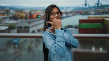 Gözlüklü ve mavi gömlekli genç Latin kadın, renkli kaplar ve bulanık bir okyanus manzarası ile dolu, yoğun bir deniz kenarı arka planında başparmak hareketleriyle neşeli poz veriyor..