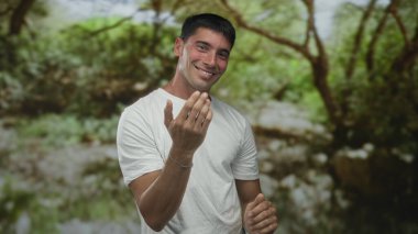 Beyaz tişörtlü adam, yemyeşil parktaki kameraya sağ eliyle işaret ediyor; arkadaşlık..