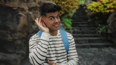 Genç adam dışarıda kulağına gülümsüyor ve el kol hareketi yapıyor. Arka planda merdivenleri olan çizgili gömlek ve sırt çantası giyiyor..