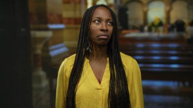 Arka planda sıra sıra sıralanmış, loş ışıklı bir kilisede sarı gömlekli bir kadın..