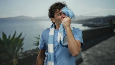 İspanyol adam güneşli bir sahilde futbol zaferini kutluyor çarpıcı bir deniz zeminine karşı çizgili bir eşarp sergiliyor tutku, heyecan ve spor ruhunu yansıtıyor..