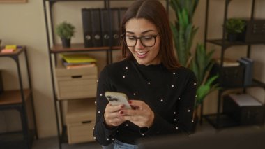 Modern ofis ortamında raflar ve bitkilerle akıllı telefon kullanan bir kadın gülümsüyor..