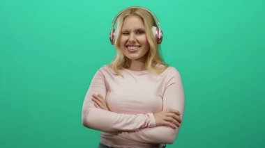 Parlak yeşil bir arka planda kulaklıkla gülümseyen, pembe bluzlu ve sarı saçlı, stüdyo ortamında neşeli davranışlar sergileyen, kollarını kavuşturmuş bir kadın..