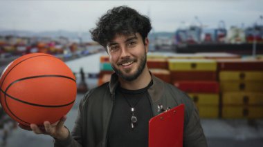 Limanda sakallı genç bir adam basketbol oynuyor. Etrafı gemi ve konteynırlarla çevrili. Günlük giysiler giyiyor ve güvenle gülümsüyor..