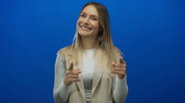 Mavi arka planda coşkuyla gülümseyen kadın, pozitifliğini ve uzun sarı saçlarla nişanını gösteriyor, parmaklarını göstererek neşeli ve canlı bir atmosfer yaratıyor..