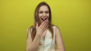 Beyaz kolsuz bluzlu, ağzı açık, parlak sarı stüdyo duvarına gülen bir kadın..