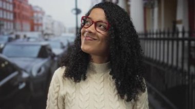 Kadın yüzü bulutlu gökyüzünün altında kırmızı gözlük ve krem süveter giyerek gülümsüyor; şehir yaşamında iyimserlik.