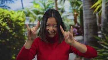 Kırmızı kazaklı genç bir kadın taştan bir jest yapıyor ve açık havada yeşil bir parkta büyük bir gülümseme sergiliyor, neşe ve neşe dolu bir enerji taşıyor..