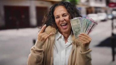 Kadın gururla Rupi banknotları gösteriyor canlı bir şehir caddesinde rock jestleri yapıyor, dışarıda neşe ve güveni ifade ediyor.