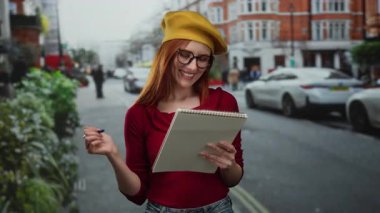 Şehir merkezinde sokakta notla gülümseyen bir kadın, açık havada çizim yapan kızıl saçlı, sanatsal ilham veren canlı bir sahne..