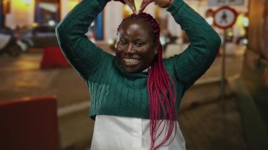 Kollarıyla gülümseyen ve kafasında kalp şekli oluşturan bir kadın. Canlı bir şehir sokak ortamında..