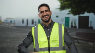 Şehir caddesinde yansıtıcı yelek giyen genç adam dışarıda yüzünü gizliyor. Binalar ve ağaçlarla karışık duygular sergiliyor.