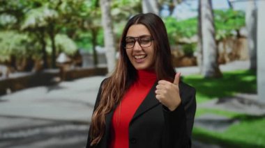 Güneşli bir parkta gülümseyen İspanyol kadın gözlüklü ve kırmızı gömlekli neşeli ve pozitif bir atmosferde ağaçlarla çevrili..