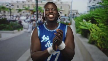 Mavi basketbol forması ve beyaz bilekliklerle kalabalık bir caddede el çırpan adam, gülümseyip geçen yayaları alkışlıyor; sevinç kutlamaları.