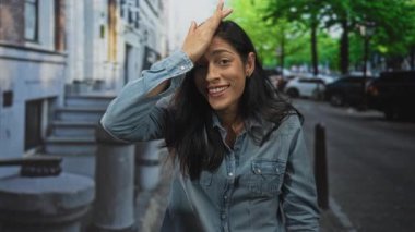 Kot gömlekli, ağzı açık, elleri alnında bir kadın bina merdivenleri ve park edilmiş bir arabanın yanında jest yapıyor..