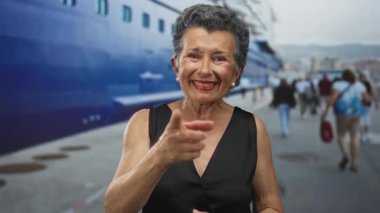 Büyük bir yolcu gemisiyle hareketli bir limanın önünde sıcak bir şekilde gülümseyen gri saçlı yaşlı bir kadın ve deniz kenarında bir neşe ve macera dolu bir şekilde yürüyen insanlar..