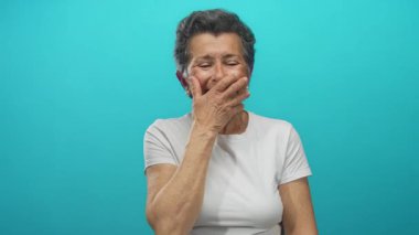 Gri saçlı yaşlı bir kadın, parlak mavi bir arka plan karşısında gülerken, ağzını kapatıyor, günlük beyaz bir tişört giyiyor ve basit bir ortamda neşe sergiliyor..