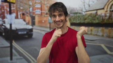 Kırmızı tişört giyen bir adam işaret parmağını şehir sokaklarında yukarı kaldırıyor; mutluluk güven ifadesi.