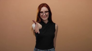 Dost canlısı bir şekilde gülümseyen bir kadın basit bir kahverengi arka plana karşı gözlüklü ve kendinden emin bir ifadeyle günlük siyah kıyafetli..