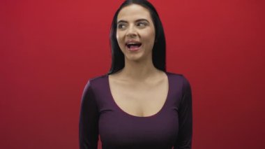 Kadın, elleri açık bir şekilde kırmızı duvara karşı gizemli bir jest yapıyor. Neşeli bir ifade ve renkli bir ortam sergiliyor..