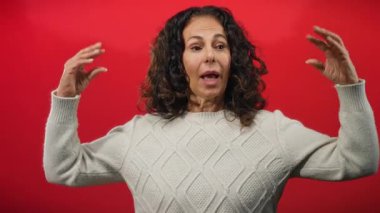 Kıvırcık saçlı İspanyol kadın jest yapıyor, kırmızı arka plan önünde süveter giyiyor..
