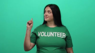 Yeşil arka plan üzerinde duran yeşil tişörtlü gönüllü kadın güven içinde yukarıya doğru bakan düşünceli bir ifade ile onaylama ve motivasyon göstermektedir.
