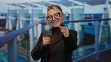 Yolcu gemisinde elinde mikrofonla el kol hareketi yapan bir kadın, açık hava arkaplanına yaslanmış, muhabir teması, davet edici ifadeler ve profesyonel kıyafetlerle somutlaştırılmış..