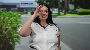 Uzun saçlı genç bir kadın gözünün üstüne iyi bir jest yapıyor, yeşil bir sokakta gülümsüyor ve geçmişi bulanık..