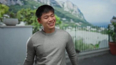 Genç adam yeşil dağ manzaralı canlı bir şehir caddesinde gülümsüyor, gri bir süveter giyiyor ve mutluluk ve pozitifliği ifade ediyor..