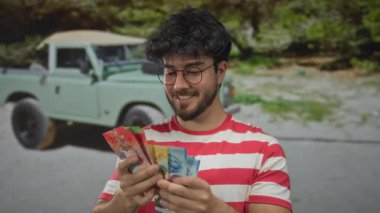 Sakallı genç adam elinde renkli İsviçre frangı banknotları tutarken arka planda klasik bir aracın olduğu yeşil bir park ayarında gülümsüyor..