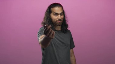 Pembe stüdyoda parmak kalp hareketi yapan ve gülümseyen, gri t-shirt giyen bir adam; eğlenceli bir davet.