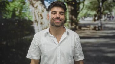 Beyaz tişörtlü genç İspanyol adam, yeşil ormandaki benekli güneş ışığının ortasında elleri ile gözleri gölgeliyor ve gülümsüyor; iyimserlik.