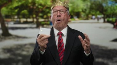 Takım elbiseli kıdemli bir adam parkta gülüyor, elinde kredi kartıyla yeşil ağaçlar ve güneş ışığı olan bir arka planda neşe saçıyor..