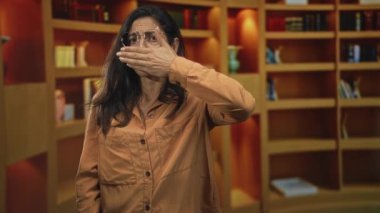 Elle gözlerini kapayan ve kütüphanede ahşap kitaplıklar ve görünür lamba ile alnına bastıran kadın; rahatsızlık.