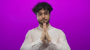 Gözlüklü ve sakallı genç adam mor arka planda gülümsüyor, açık renk gömlek giyiyor, pozitiflik ve sakin tavır takınıyor..